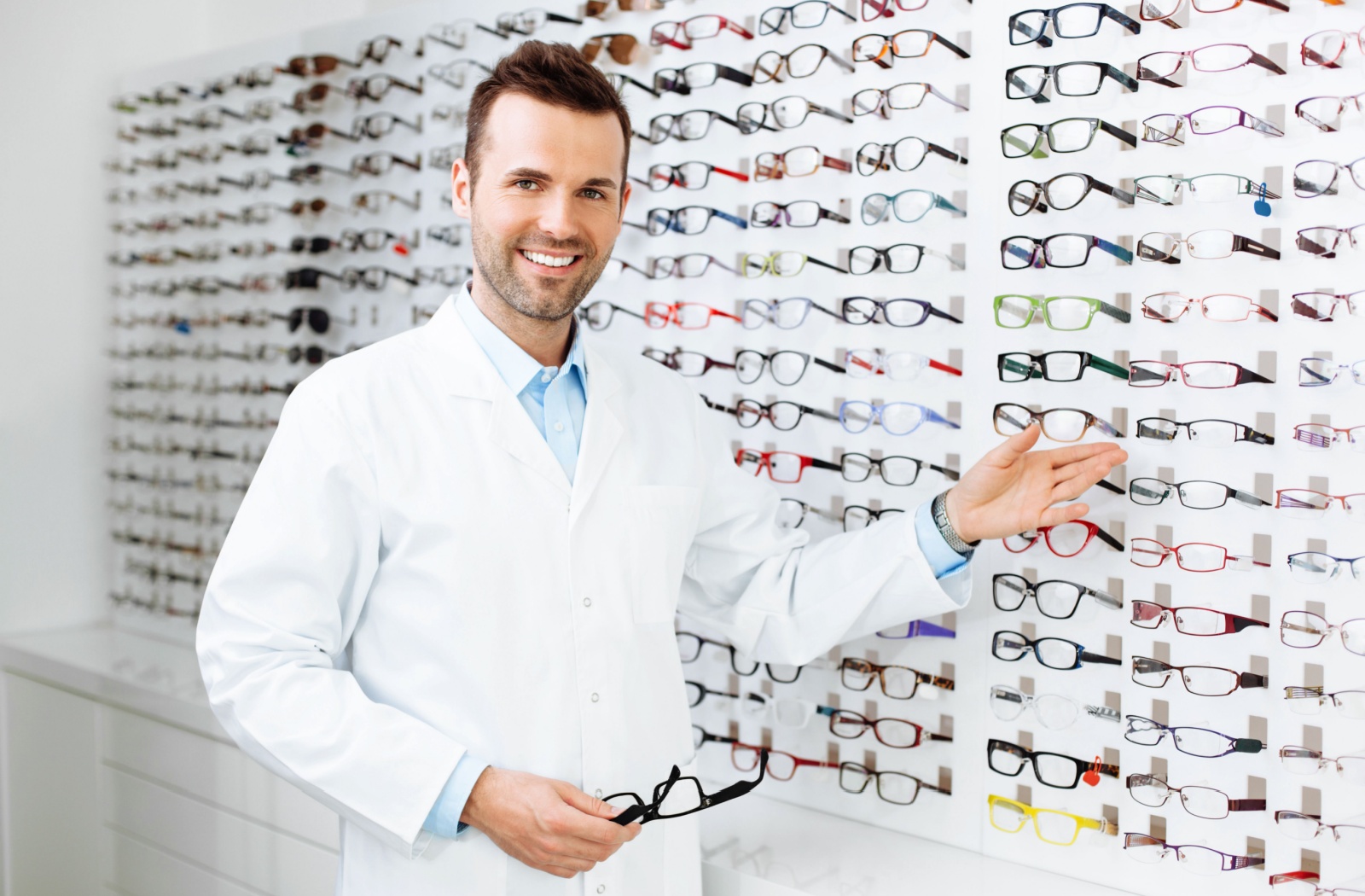 A male optometrist stands in front of a wall of eyeglasses frames that the practice is selling