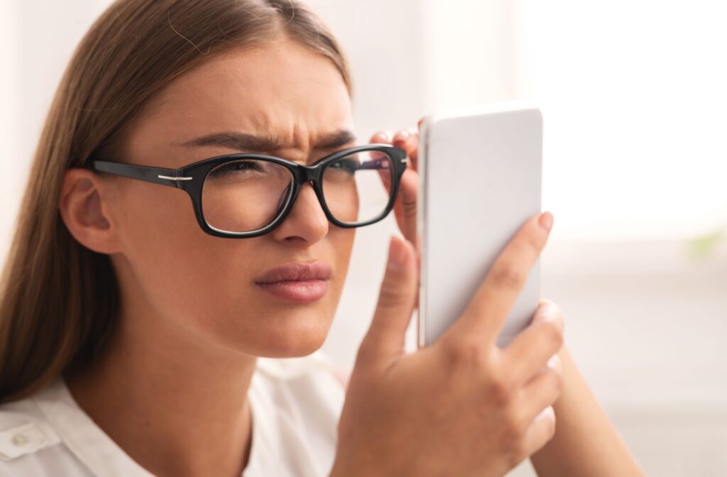 A woman wearing glasses squints at her phone while holding it close to her face.