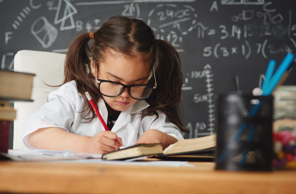 A young girl in pigtails and black wide-rimmed glasses dutifully writes on a piece of paper, surrounded by school supplies