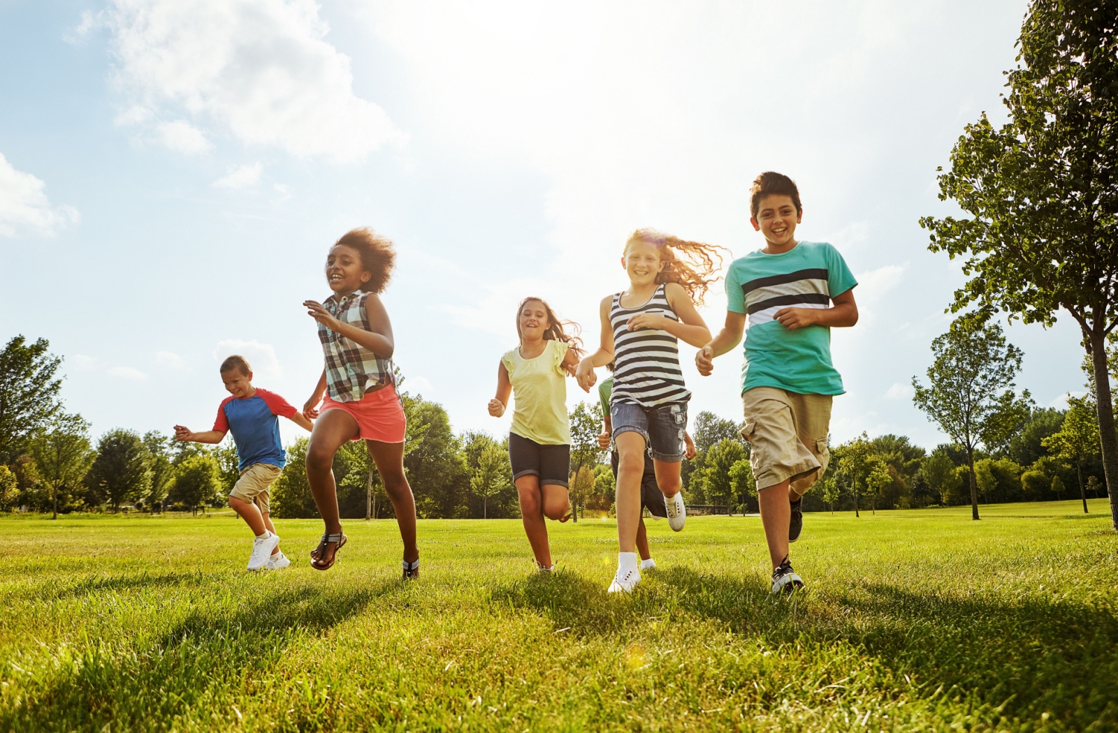 A group of several school-age children run through a green field, smiles on their faces and the sun at their backs.