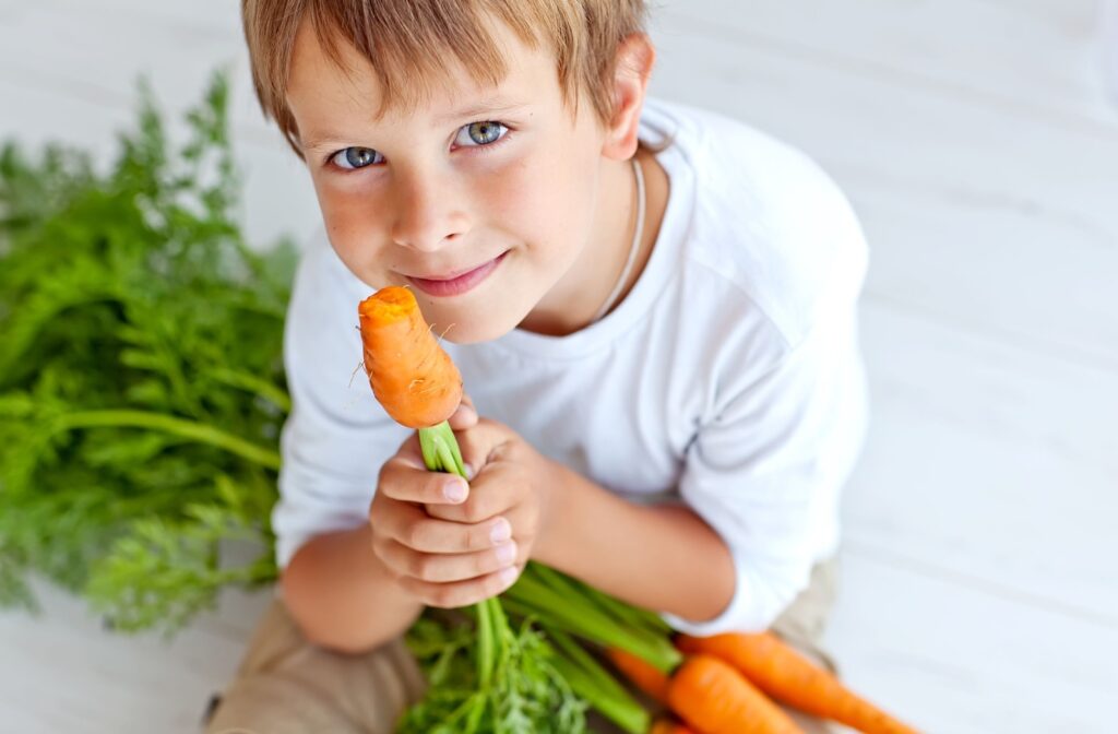 A child holds half a carrot in their hands and smiles with a bundle of carrots in their lap.