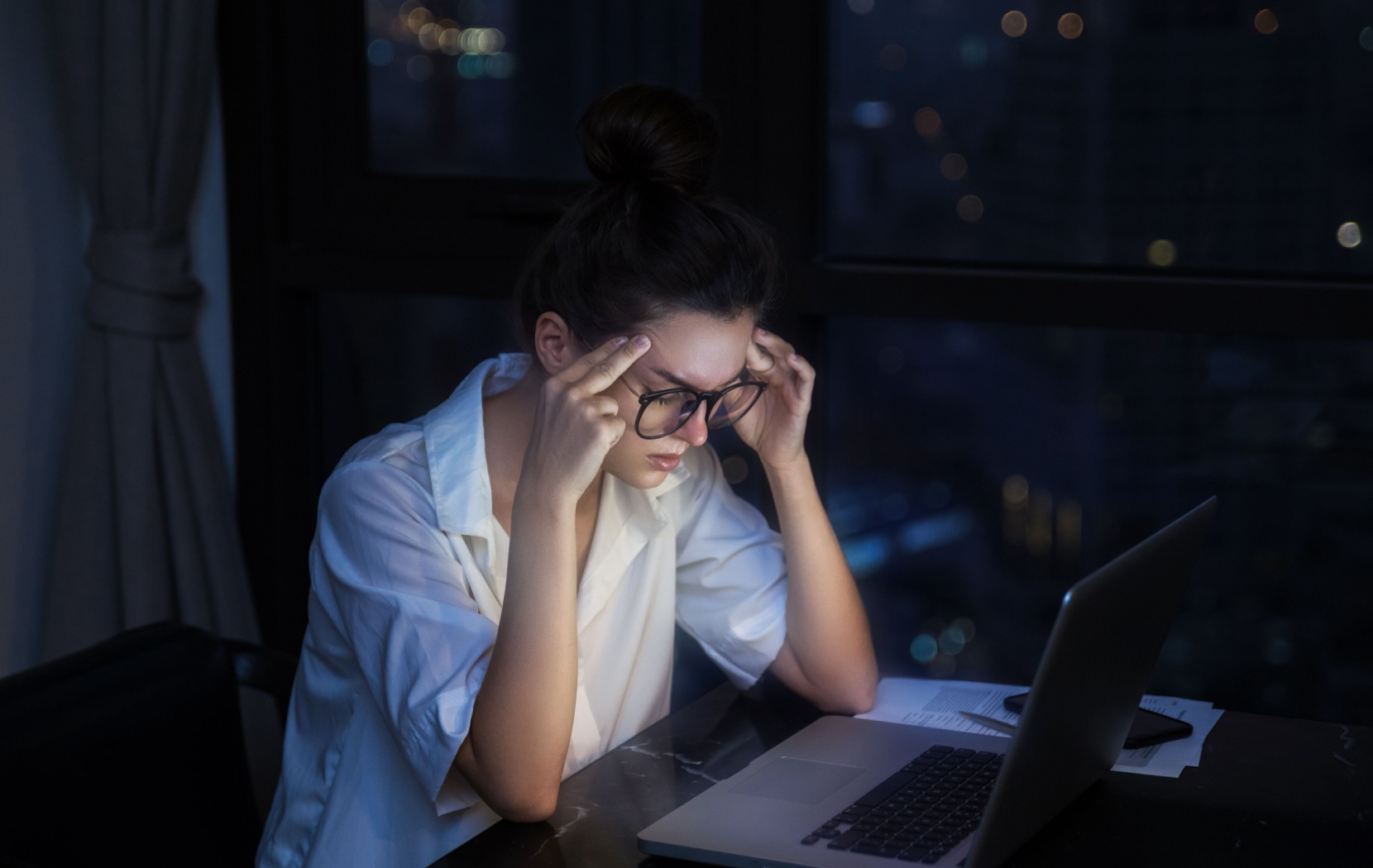 A young adult rubs their temples and squints their eyes as the light of a computer screen lights up their face.